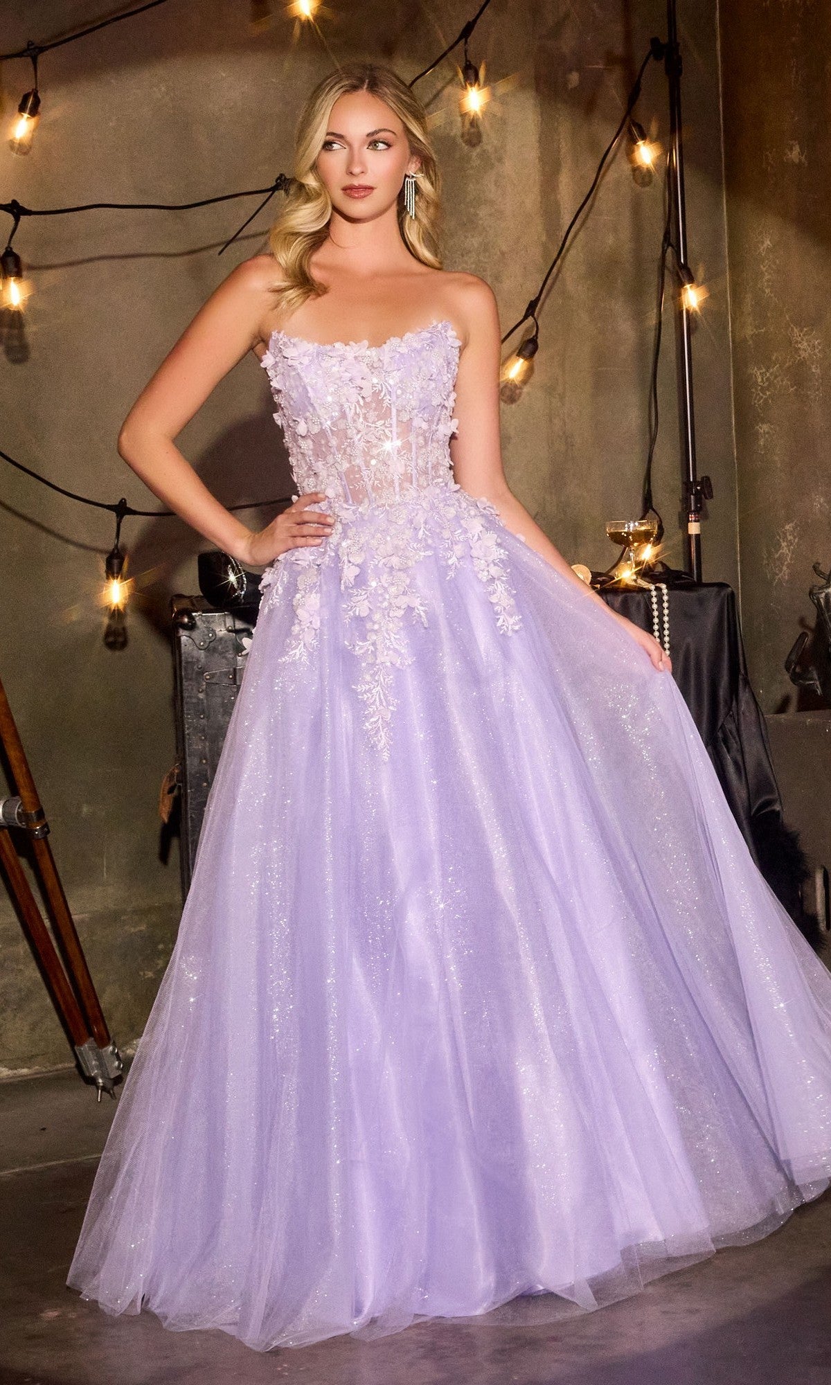 A woman wears the Ladivine C174 Long Prom Dress in lavender, featuring a strapless tulle design with 3D floral embroidery and a sheer corset bodice, standing indoors amid string lights and industrial decor.